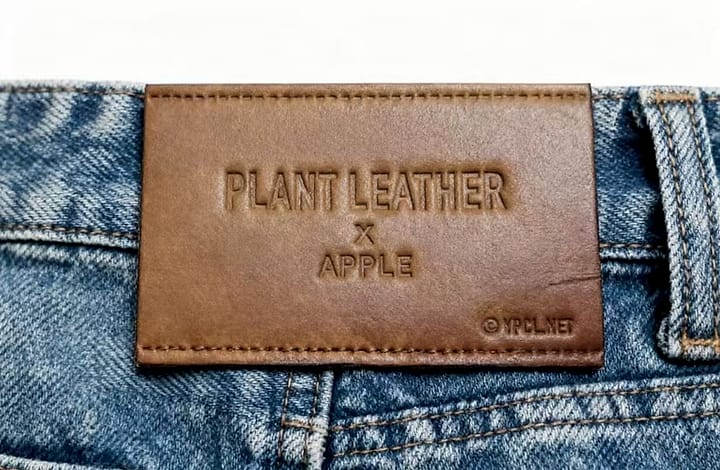 Plant leather patch photographed — PLANT LEATHER X APPLE and the YPCLYET copyright mark deep-embossed, with double dark-brown reinforced stitching, on an eco-concept denim back waist