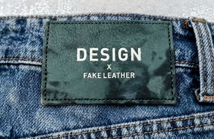 PU faux leather patch photographed — screen-printed and hot-foil lettering in a clean sans-serif, on a designer-collab denim back waist