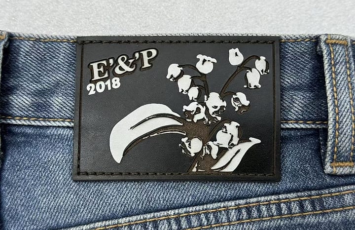 Relief leather patch photographed — black plain-grain cowhide with laser-engraved, colour-filled relief and crisp edges, on a brand anniversary denim back waist